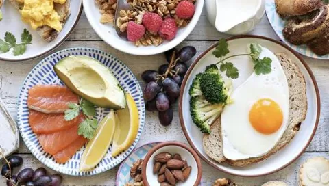 Ovanifrån dukat bord med stekt ägg på bröd, avokado och lax, granola med hallon, druvor och nötter.