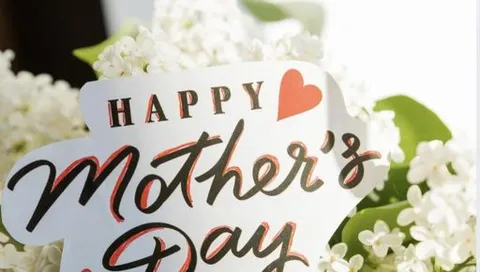 Närbild av ett kort med texten “Happy Mother’s Day” och ett hjärta, omgiven av vita blommor och gröna blad.