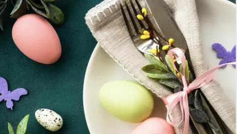 Tallrik med bestick i linneservett med rosa band och kvistar, omgiven av pastellfärgade påskägg och dekorationer.