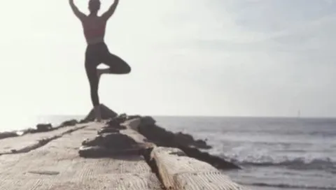 En silhuett av en person gör yogaposen träd på en träbrygga vid havet, med vågor och ljus himmel i bakgrunden.